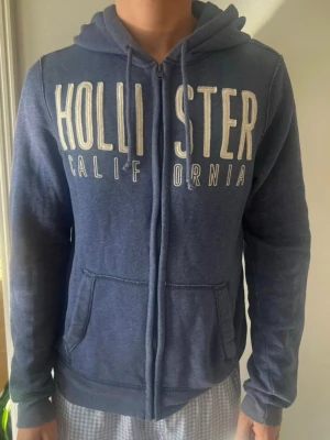 Blå zip hoodie från Hollister - Snygg blå hoodie från Hollister med dragkedja framtill och stora vita bokstäver på bröstet. Klassisk huva och två fickor framtill. Tillverkad i mjukt material som känns skönt mot huden. Perfekt för en avslappnad och cool stil.