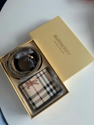 Rutigt plånbok från Burberry - Snygg plånbok från Burberry med klassiskt beige rutmönster och detaljer i svart och vinrött. Plånboken har en elegant design med Burberrys ikoniska mönster och ett tryckt motiv på framsidan. Materialet är strukturerad canvas.