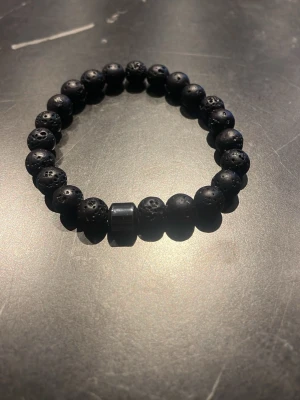 Svart lavastenarmband med pärlor - Snyggt armband med runda svarta lavastenpärlor och en större metallisk pärla i mitten. Elastiskt band som gör det enkelt att ta på och av. Perfekt accessoar för en stilren och modern look.