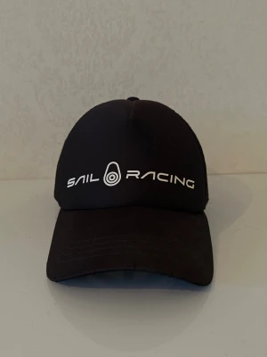 Svart keps från Sail Racing - Snygg svart keps från Sail Racing med vit logga och text framtill. Justerbar baktill för perfekt passform. Perfekt accessoar för en sportig och avslappnad stil.