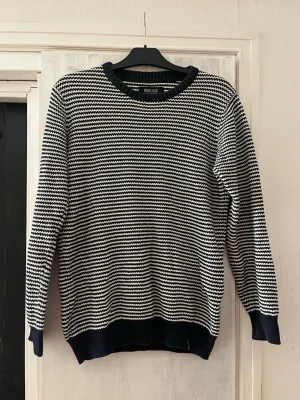 Randig stickad tröja  - Snygg stickad tröja med smala svarta och vita ränder. Tröjan har rund halsringning, långa ärmar och ribbade muddar vid ärmslut och nederkant. Perfekt för dig som gillar klassisk och enkel stil. 