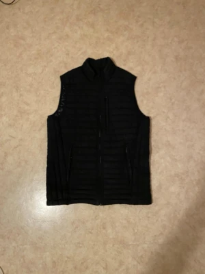 Svart dunväst med dragkedja - Säljer en svart dunväst med hög krage och dragkedja framtill. Västen har horisontella quiltade sömmar och flera fickor med dragkedja. Perfekt för lager-på-lager och ett stilrent val till hösten och vintern.