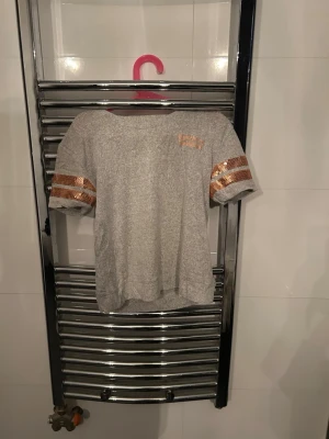 Grå t-shirt med paljettdetaljer - Snygg grå t-shirt från Levi’s med korta ärmar och två rader kopparfärgade paljetter på ärmarna. Liten levi’s-logga i paljetter på bröstet. Perfekt för dig som gillar lite extra bling i vardagen.