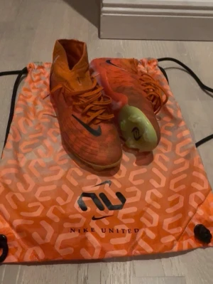Nike phantom luna elite fg united orange fotbollsskor - Säljer ett par Nike phantom luna och elite fg united fotbollsskor i en snygg orange färg med svarta detaljer och högt skaft. Skorna har snörning och är tillverkade i syntetmaterial för bra grepp och känsla på planen. Medföljer matchande orange Nike United-påse.