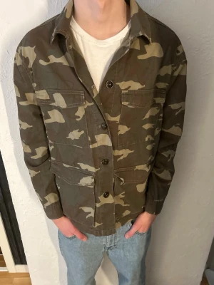 Camo overshirt med fickor - Säljer en snygg overshirt i camouflage-mönster med fyra stora fickor framtill och knäppning med knappar. Jackan är i bomullstyg och har klassisk krage. Perfekt för lager-på-lager och streetwear-stil.