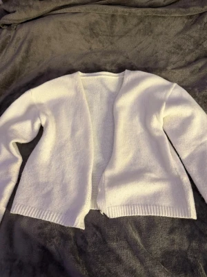 Vit stickad kofta med öppen front - Säljer en enkel och stilren vit stickad kofta med öppen front och långa ärmar. Koftan har ribbade muddar vid ärmslut och nederkant, perfekt att slänga över en t-shirt eller topp. Materialet är mjukt och bekvämt.