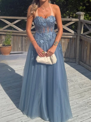 Balklänning dressroom  - Säljer en magisk blå axelbandslös balklänning med blommiga spetsdetaljer och transparenta partier upptill. Kjolen är i flera lager tyll för en extra drömmig look. Perfekt för bal eller student! Kontakta mig för mer information om storlek, kvitto mm. Uppsydd till min längd dvs 172cm och jag har dessutom väldigt långa ben💕💕 endast använd en gång. Det medföljer även armpuffar som går att välja om man vill använda eller inte🤗