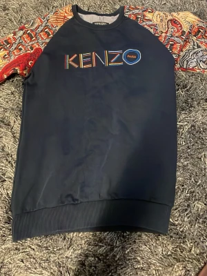Kenzo sweatshirt med mönstrade ärmar - Säljer en mörkblå Kenzo sweatshirt med färgglatt broderat Kenzo Paris-tryck på bröstet. Ärmarna har ett unikt mönster med vargar och röda detaljer som verkligen sticker ut. Tröjan har rund halsringning och ribbade muddar. Perfekt för dig som vill ha något som syns!