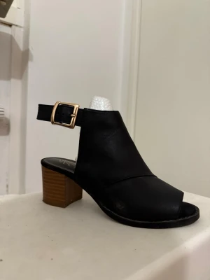 Svarta sandaler med klack från SO - Stilrena svarta sandaler från SO med öppen tå och bred rem över foten. Sandalen har en chunky träfärgad klack och justerbart spänne i guld. Tillverkad i konstläder med modern design som passar till många looks.