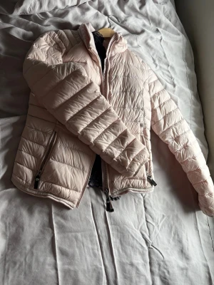 Ljusrosa pufferjacka från Cedrico - Supersnygg ljusrosa pufferjacka från Cedrico med quiltad design och hög krage. Jackan har dragkedja framtill, två sidofickor med dragkedja och mjukt innerfoder. Perfekt för kalla dagar och ger en clean, modern look.