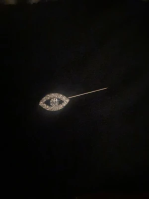 Gnistrande broschnål med strass - Säljer en elegant broschnål i silverfärgad metall med ögonformad design. Dekorerad med klara strass-stenar runtom och en större sten i mitten som ger extra bling. Perfekt accessoar för att lyfta din outfit med lite extra glitter.