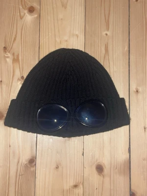 Svart mössa från C.P. Company - Säljer en svart stickad mössa från C.P. Company med de ikoniska inbyggda goggles på uppviket. Mössan är tillverkad i mjukt material och har en modern look som sticker ut. Perfekt accessoar för dig som gillar unik stil.