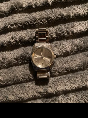 Klassisk silverfärgad metallklocka - Stilren klocka i silver med rund urtavla och metallarmband. Urtavlan har minimalistiska markeringar och en räfflad kant runt glaset. Perfekt accessoar för dig som gillar en clean och tidlös look.