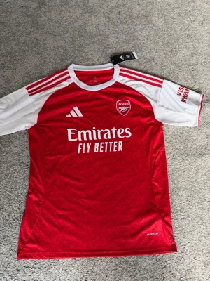Arsenal matchtröja röd/vit Adidas - Snygg Arsenal matchtröja från Adidas i rött och vitt med klubbmärke och sponsortryck på bröstet. Tröjan har vita raglanärmar med röda detaljer och Adidas klassiska ränder på axlarna. Tillverkad i lätt och ventilerande funktionsmaterial. Helt ny