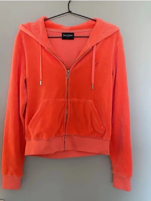 Juicy Couture orange velour hoodie - Juicy Couture hoodie i orange velour med dragkedja framtill, huva och fickor. Klassisk logga broderad på bröstet och ribbade muddar vid ärmslut och nederkant. Perfekt för en avslappnad och trendig look.