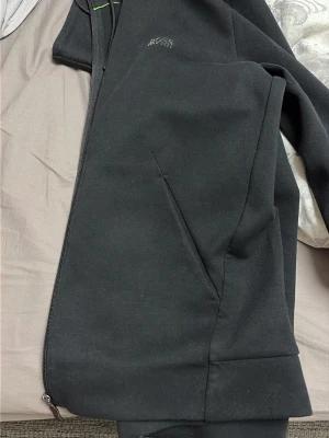 Svart hoodie från Hugo Boss - Snygg svart hoodie från Hugo Boss med dragkedja framtill och diskret logga på bröstet. Hoodien har två sidofickor och är tillverkad i mjukt bomullsmaterial. Perfekt för en avslappnad och stilren look.