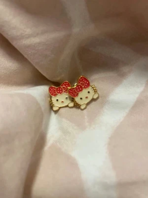 Hello Kitty örhängen med röd rosett - Säljer ett par söta Hello Kitty örhängen i guldtonad metall med vit och röd emalj. Örhängena har motiv av Hello Kittys ansikte med en röd prickig rosett och små guldfärgade detaljer. Perfekta för dig som gillar kawaii-stil och vill ha något unikt i smyckessamlingen.