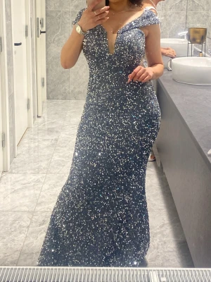 Glittrig mörkblå offshoulder klänning - Säljer en lång, glittrig kall grå elegant klänning med offshoulder och djup v-ringning. Klänningen har en figurnära passform och är täckt med paljetter som ger ett riktigt lyxigt intryck. Perfekt för bal eller fest! ✨ sidorna har samma öppning som framsidan (som vid bröstet), passar någon som är 160-170. Haft den på mig 1 gång i 4 timmar då jag aldrig går på fester. Ordninarie pris var 3500 kr 