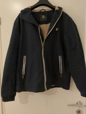 Marinblå vindjacka från Lyle & Scott - Stilren marinblå vindjacka från Lyle & Scott med dragkedja framtill, två vita dragkedjeförsedda fickor och beige innerfoder. Sparsamt använd, inga defekter. Lite skitig men kan tvätta innan jag skickar. Pris går att diskutera.