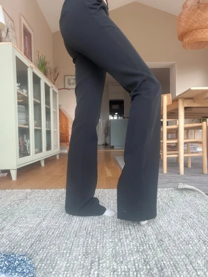 Svarta utsvängda kostymbyxor med låg midja - Svarta kostymbyxor med utsvängda ben och låg midja. Klassisk design med hällor för skärp och dold dragkedja framtill. Perfekta för en stilren och trendig look. Är lite långa på mig, är 167. 