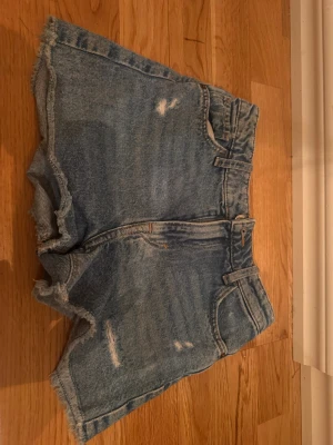 Blåa jeansshorts med rå kant - Säljer ett par blå denimshorts med hög midja och råa, fransiga kanter. Shortsen har slitna detaljer framtill och bakfickor. Perfekta för varma dagar och en avslappnad stil.