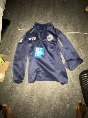 Blå träningsjacka från Umbro - Säljer en mörkblå träningsjacka från Umbro med halv dragkedja och tryckta loggor på bröstet och ryggen. Jackan har klubbmärke, sponsorloggor och namnet 'Mugisa' på ryggen. Perfekt för fotbollsträning eller sportaktiviteter.