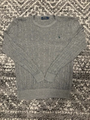 Grå kabelstickad tröja Polo Ralph Lauren - Kabelstickad grå tröja från Polo Ralph Lauren med rund halsringning och diskret broderad logga på bröstet. Långa ärmar och ribbade muddar vid ärmslut och nederkant. Klassisk och stilren design som passar till det mesta. Tröjan är i storlek S men passar även i storlek M. Hör av er vid eventuell frågor eller funderingar!!