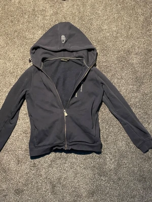 Mörkblå hoodie från Sail Racing - Mörkblå hoodie från Sail Racing med dragkedja och broderad logga på huvan. Mjuk insida i fleece och smidig passform. Perfekt för dig som gillar stilren och sportig look. Klassisk modell med långa ärmar och fickor framtill.