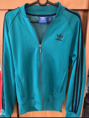 Turkos Adidas hoodie med dragkedja - Snygg turkos hoodie från Adidas med dragkedja framtill och svarta klassiska ränder längs ärmarna. Tröjan har två fickor, ribbade muddar och Adidas logga på bröstet. Perfekt för en sportig och avslappnad stil.