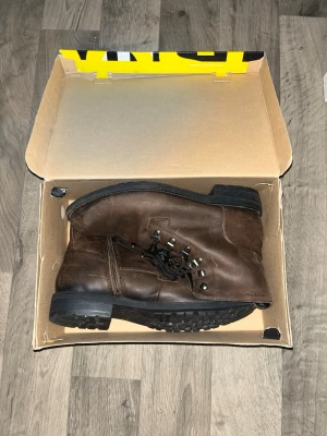 Bruna läderkängor med grov sula - Säljer ett par bruna kängor i läder med robust svart sula och metallhål för snörning. Kängorna har klassisk design med högt skaft och svarta snören, perfekta för dig som vill ha en stilren och tålig sko till hösten och vintern.