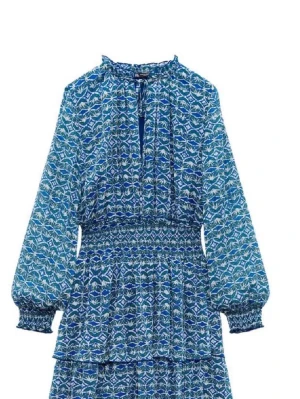 Blå mönstrad klänning med volanger - Blå klänning med vitt och grönt mönster, långa ärmar och volangdetaljer. Klänningen har smock i midjan och vid ärmsluten samt knytning i halsen. Perfekt för dig som gillar bohemisk stil och vill ha något unikt.