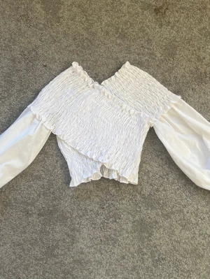 Vit omlottopp med volangdetaljer - Superfin vit omlottopp med smockad struktur och volangkant. Toppen har långa ärmar i glansigt material och en snygg v-ringning. Perfekt att styla med jeans eller kjol för en trendig look.