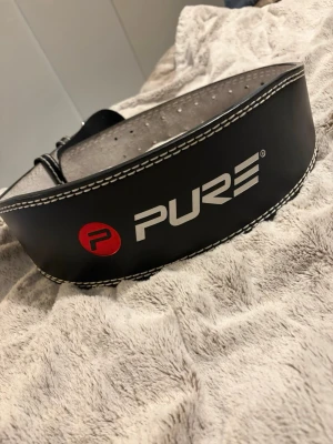 Svart träningsbälte från PURE - Säljer ett svart träningsbälte från PURE med vit kontrastsöm och logga i vitt och rött, perfekt för gymmet när du vill ha extra stöd vid tunga lyft. Justerbar storlek med flera hål och rejäl spänne.