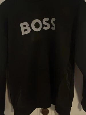 Svart sweatshirt från BOSS - Svart sweatshirt från BOSS med stor vit logga på bröstet. Klassisk rund halsringning och lång ärm. Perfekt för en clean och stilren look. Mjuk och bekväm, passar till det mesta i garderoben.