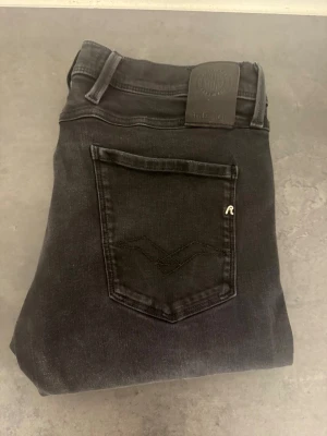 Svarta Replay Anbass slim jeans stl 33/32 - Snygga svarta slim fit jeans från Replay med klassisk femficksmodell och diskret logga på bakfickan. Jeansen har en smal passform och är tillverkade i stretchigt denimtyg för extra komfort. Perfekta till en avslappnad streetwear-look.