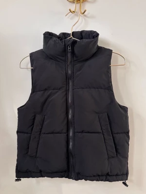 Svart dunväst med hög krage - Stilren svart dunväst med hög krage och dragkedja framtill. Västen har två snedställda fickor och quiltad design som ger en puffig look. Perfekt att bära över en hoodie eller tröja för extra värme. Enkel och tidlös modell som passar till det mesta och perfekt till vår och höst.