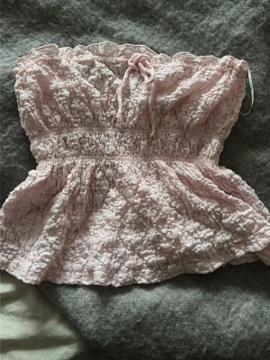 Ljusrosa bandeau topp Gina Tricot - Supersöt ljusrosa bandeau topp från Gina Tricot med volangkant upptill och smockad midja. Toppen har spetsdetaljer och en liten rosett framtill. Perfekt för sommarens alla tillfällen och ger en romantisk vibe.