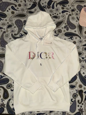 Vit hoodie från CD - Snygg vit hoodie från CD med stor färgglad logga dekorerad med blommor på bröstet. Klassisk huva med dragsko och magficka framtill. Tillverkad i mjukt bomullsmaterial som känns skönt mot huden. Perfekt för dig som vill ha en stilren men ändå unik look.