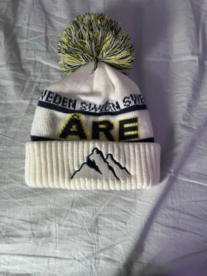 Vit Åre mössa med tofs och brodyr - Säljer en Åre mössa för att jag aldrig använder den längre. Den har en sminkfläck på insidan men ingenting som syns när man har på den.