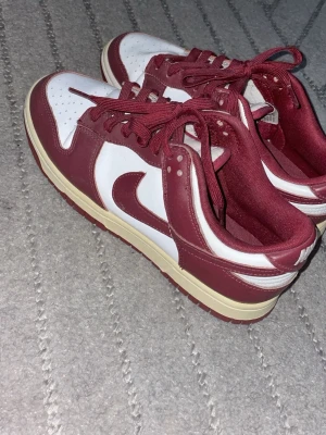 Nike Dunk Low röd/vit sneakers - Säljer ett par Nike Dunk Low sneakers i rött och vitt. Skorna har klassisk låg siluett, röda snören och detaljer samt vit tåbox och sidor. Yttermaterialet är slätt skinn och sulan är beige med röd undersida. Perfekta för dig som gillar streetwear och snygg design.