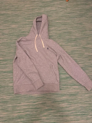 Grå hoodie från Polo Ralph Lauren - Snygg grå hoodie från Polo Ralph Lauren med klassisk broderad logga på bröstet. Hoodien har vita dragsnören, känguruficka och ribbade muddar. Tillverkad i mjuk bomullsblandning för skön känsla. Perfekt för en avslappnad och stilren look. Passar dig runt 175 cm