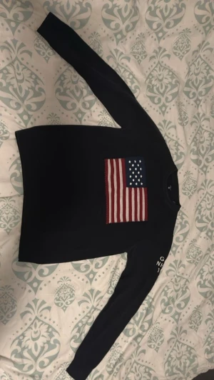 Svart tröja med USA-flagga från Gant -  svart långärmad tröja från Gant med stor amerikansk flagga broderad på bröstet. På ena ärmen står det 'GANT NH-CT 1949' i vitt. Storlek M men de kan passa Storlek S.
