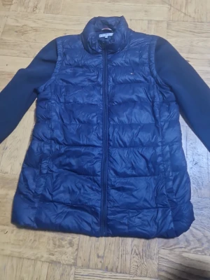 Marinblå dunväst från Tommy Hilfiger - Snygg marinblå dunväst från Tommy Hilfiger med quiltad design och diskret logga på bröstet. Västen har hög krage och dragkedja framtill. Perfekt för lager-på-lager och håller dig varm med sin lätta dunfyllning.