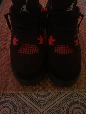 Air Jordan 4 svart och röd sneakers - Säljer ett par Air Jordan 4 sneakers i svart och rött med klassisk mesh på ovandelen och coola detaljer. Skorna har högt skaft, ikonisk Jumpman-logga på plösen och en robust sula med synlig Air-enhet. Perfekta för dig som vill sticka ut med stil.
