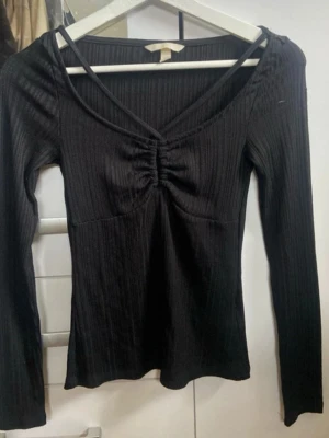 Svart ribbad långärmad topp - Snygg svart ribbad topp med lång ärm och v-ringning. Toppen har en smickrande rynkning framtill vid bysten och är tillverkad i ett stretchigt material som sitter tajt mot kroppen. Perfekt att styla med jeans eller kjol för en trendig look.