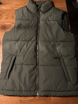 Mörkgrön väst med dragkedja - Stilren mörkgrön väst med hög krage och två fickor framtill och stängs med dragkedja. Perfekt nu till våren