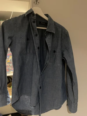 Blå overshirt från Polo Ralph Lauren - Snygg mörkblå overshirt från Polo Ralph Lauren i bomull. Klassisk skjortkrage, två bröstfickor med knappar och långa ärmar. Perfekt lager-på-lager-plagg med avslappnad passform och stilrena detaljer. Passar 160-165