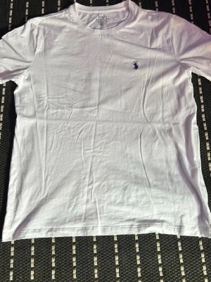 Vit t-shirt från Polo Ralph Lauren - Vit t-shirt från Polo Ralph Lauren med klassisk rund hals och diskret broderad logga på bröstet. Modellen är slim fit och tillverkad i mjuk bomull, perfekt för en clean och stilren look.