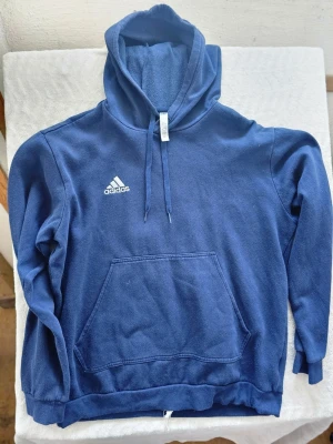 Blå hoodie från Adidas med huva - Mörkblå hoodie från Adidas med klassisk huva och dragsko. Stor känguruficka framtill och Adidas-logga i vitt på bröstet. Tillverkad i mjuk bomullsblandning som känns skön mot huden. Perfekt för en avslappnad och sportig stil.
