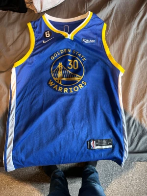 Golden State Warriors basketlinne Nike - Snyggt blått Golden State Warriors basketlinne från Nike med gula och vita detaljer. Nummer 30 och lagets logga på bröstet. Tillverkat i ventilerande meshmaterial, perfekt för basket eller streetwear. NBA-märke och etikett med storlek 52 på nederkanten.